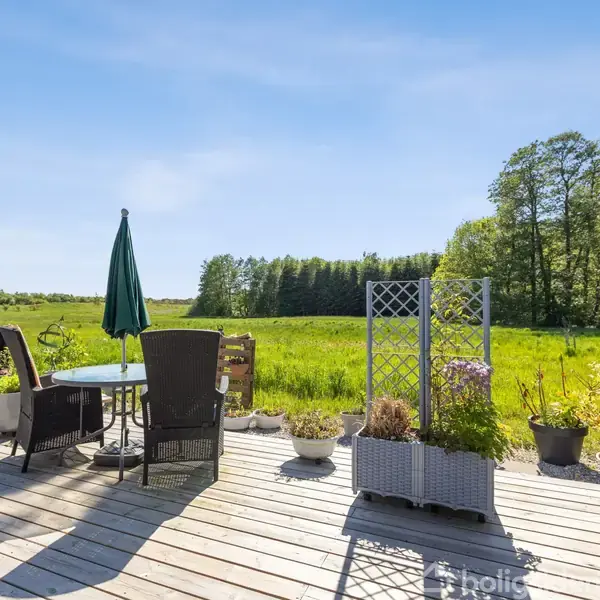 En træterrasse med et bord, parasol og stole samt blomster i krukker. Det hele vender ud mod en grøn mark med træer i baggrunden under en klar blå himmel.