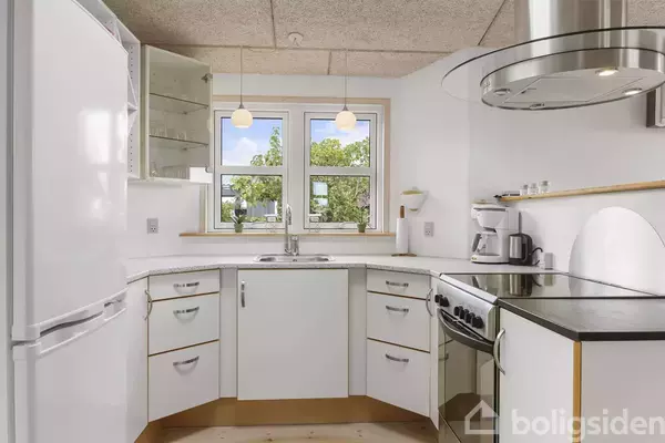 Køkken med hvide skabe og apparater indbyggede. Køleskab til venstre og ovn med gaskomfur til højre. To vinduer med udsigt til træer. Loftlamper hænger over vasken.