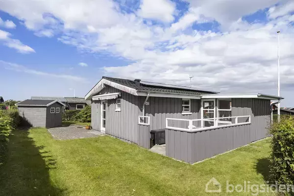 Et gråt træhus står alene, omgivet af en velplejet græsplæne og en mindre udbygning. Huset har hvide vinduer og en veranda, med blå himmel og skyer i baggrunden.