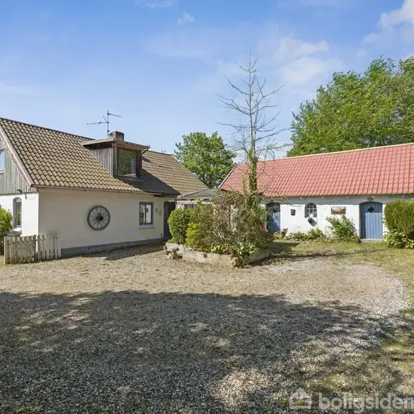 Et hvidt bondehus med rødt tag står til venstre omgivet af en gårdsplads med grus. En tilstødende bygning med blå døre og træer omkring. Solrig dag, grøn natur bagved.