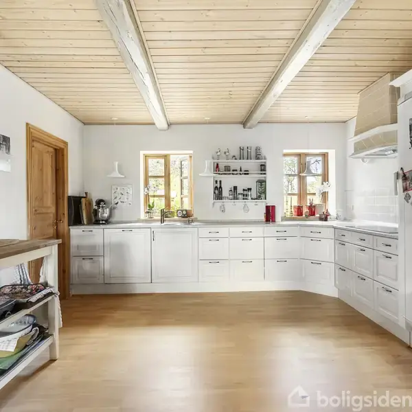 Et lyst køkken med hvide elementer og træloft. Trævinduer slipper naturligt lys ind over de vinklede køkkenskabe. Til venstre står et lille bord med hylder.