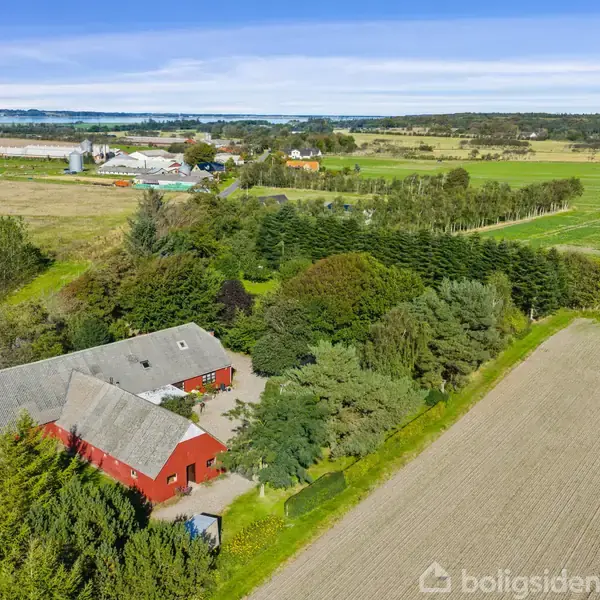 En gård med røde bygninger ligger blandt grønne træer, omgivet af åbne marker og landlige landskaber under en blå himmel.