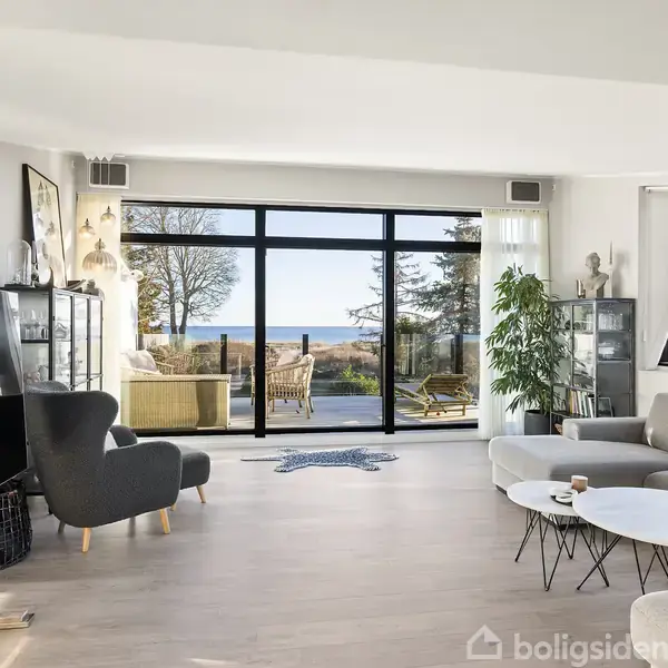 En moderne stue med en grå sofa og to stole omkring et sæt runde borde. Store vinduer viser en terrasse med udsigt til havet og træer udenfor.