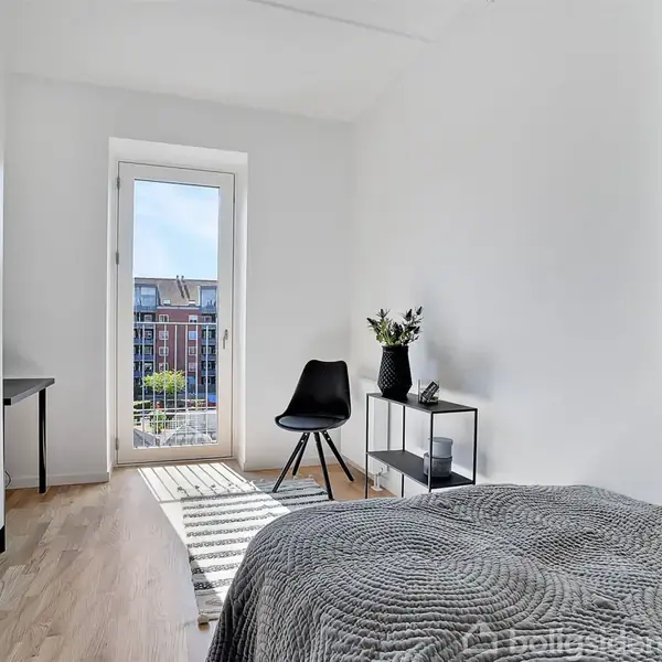Et lyst soveværelse med en seng dækket af et gråt tæppe. Et lille bord med planter og en stol står ved glasdøren til en lille altan. Kigger ud over bytage.