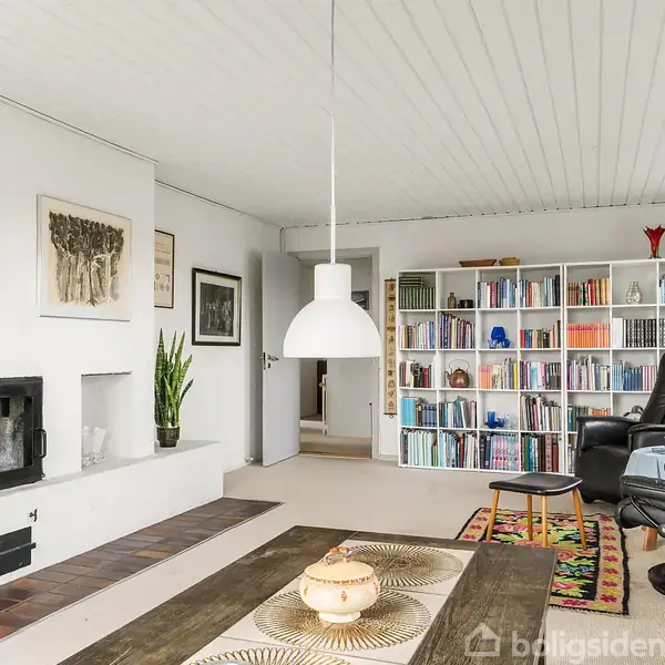 En stue med et sort lænestolssæt, en bogreol fyldt med bøger, en væghængt pejs og en loftlampe. Væggene er pyntet med billeder og et tæppe ligger på gulvet.