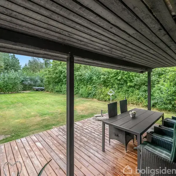 Veranda med sort træbord og fire flettede stole står under et overdækket træloft. Udsigten viser en stor græsplæne omgivet af tætte, grønne buske og træer.