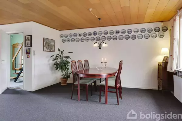 Et rundt spisebord med seks stole står i et rum med en grå gulvtæppe. En plante i krukke står til venstre, og på væggen hænger mange runde billeder.