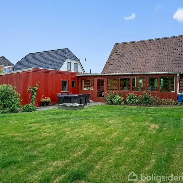 Rødt hus med tilstødende terrasse og havemøbler, beliggende i frodig have med græsplæne, buske og havepynt i form af en lille vindmølle. Baggrunden er blå himmel med få skyer.