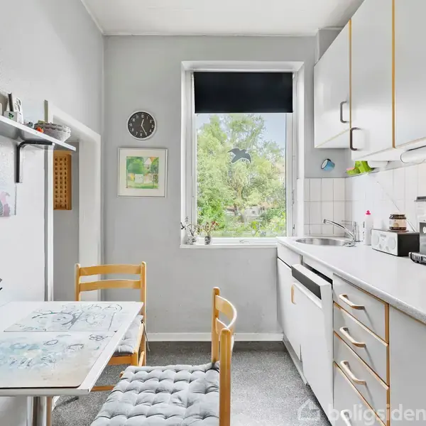 Bord med to stole står ved væg med hylder, mens kaffemaskine er på køkkenbord under skabe. Vinduet viser grøn have. Uret og maleri hænger på væggen.