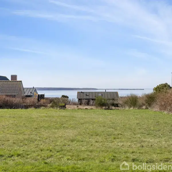Hus ved siden af græsplæne står stille, med udsigt over havet i baggrunden, omgivet af buske og himmel med lette skyer.