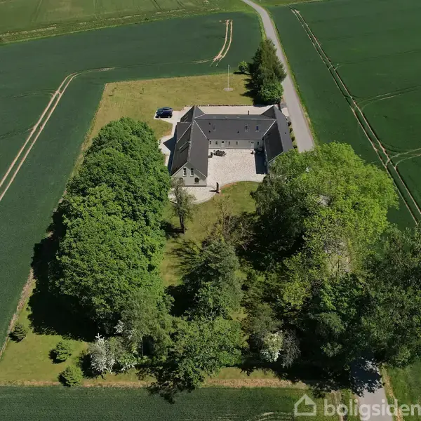 En stor gård bygning står stille i et åbent landbrugsområde, omgivet af grønne marker og træer. En enkelt bil er parkeret ved gården.