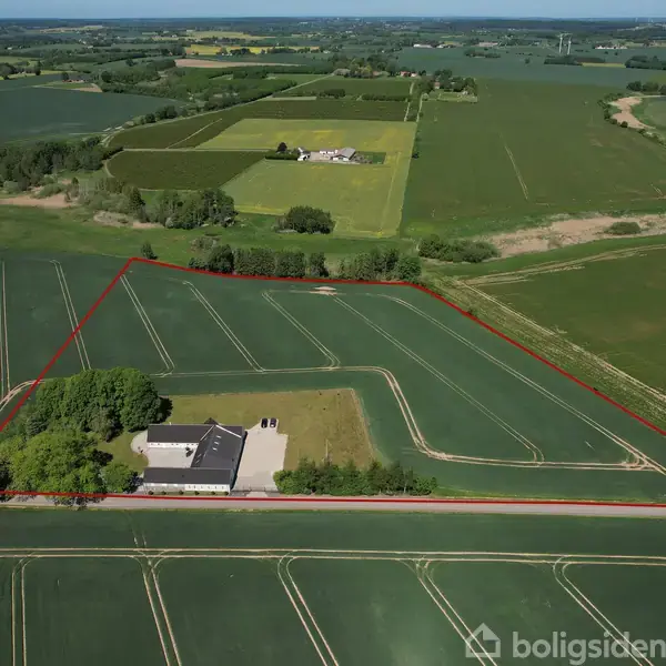En hvid bygning ligger centralt i et stort grønt markområde med marker, trærækker og en vej foran. Et rødt omrids indikerer ejendomsgrænser.