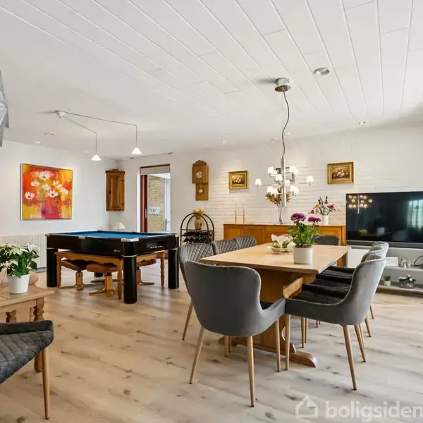 Et stort opholdsrum med et spisebord omringet af stole. Et billardbord står i baggrunden. Rummet har lyst trægulv, loftlamper og vægkunst. Et TV står i hjørnet ved vinduet.