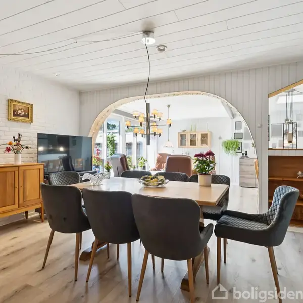 En spisestue med et stort bord omgivet af polstrede stole, dekoreret med planter og frugtskål. Rummet har trægulv og hvide vægge, med en åbning til en lys stue.