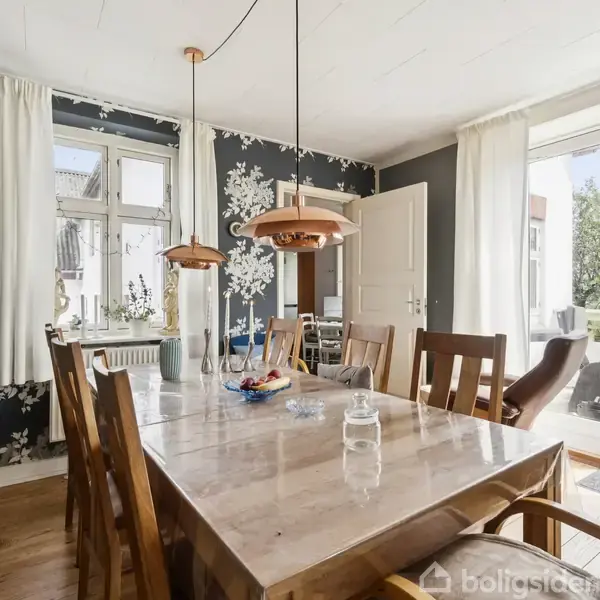 Spisebord er dækket med skål og glas, omgivet af træstole, i en stue med blomstrede tapeter. Store vinduer og døre slipper lys ind, der åbner mod en terrasse.