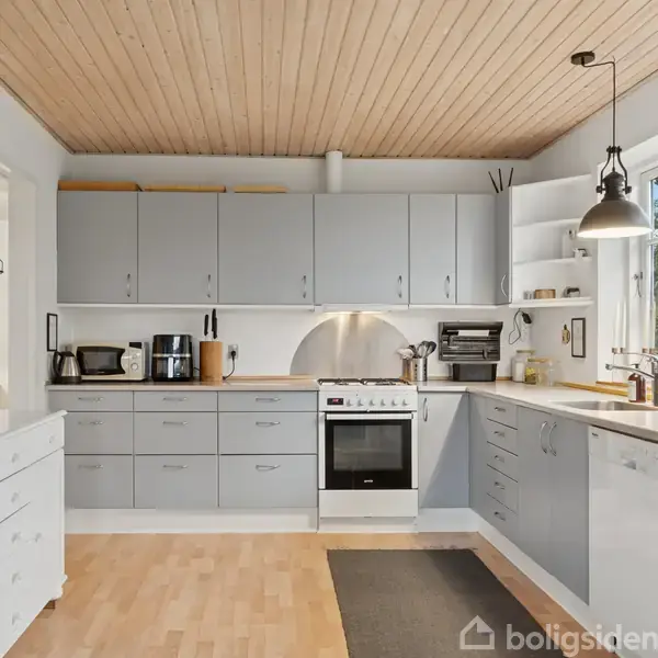 Køkken med grå skabe og skuffer, elektrisk ovn og komfur, kaffemaskine og brødrister. Bordplads omkring vask og køkkenudstyr. Træloft og lyse vægge i en moderne, minimalistisk indretning.