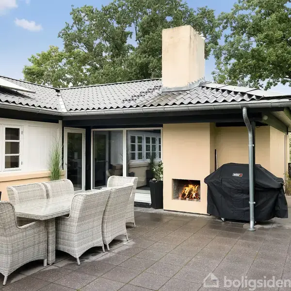 Terrasse med møbleret spisebord står foran et moderne hus. En grill og en udendørs pejs med brændende ild er til højre. Møblerne er i flet, omgivet af grønne planter.