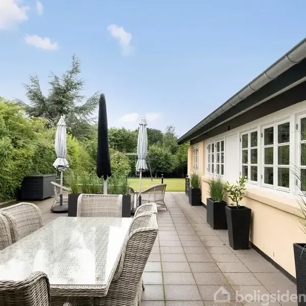Et udendørs terrasseområde med bord og stole i flet, omgivet af planter i sorte potter. Paraplyer står oprejst. Terrassen støder op til et gult hus med hvide vinduer.
