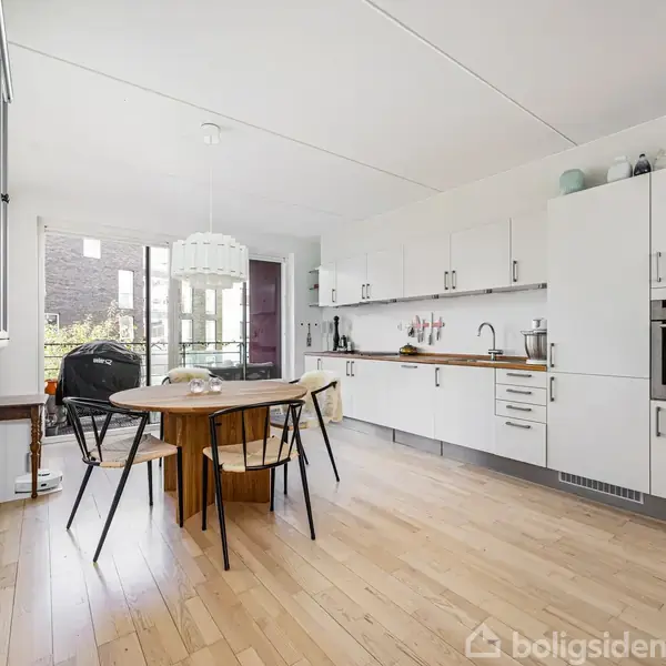 Køkken med hvidt interiør, hvor et rundt spisebord står i midten omgivet af stole. Store vinduer i baggrunden giver udsigt til en altan og bygninger.