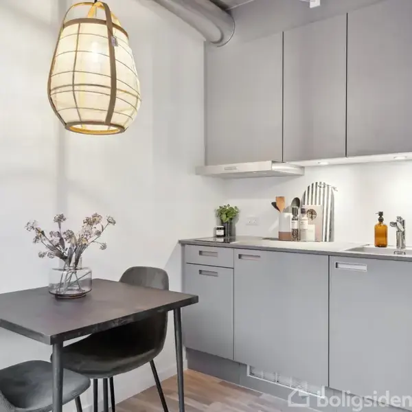 Et lille køkken med grå skabe, en vask, og forskellige køkkenredskaber. Et bord til to personer med blomster i en vase. Ovenover hænger en flettet lampe.