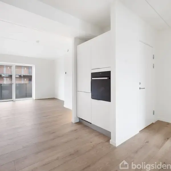 Et lyst, moderne køkken med indbygningsovn i et hvidt skab. Trægulv strækker sig mod en stue med store vinduer mod en balkon.