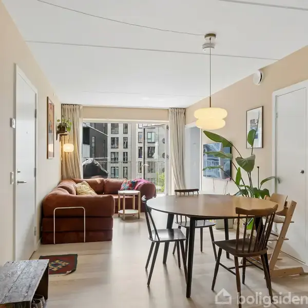 En lys stue med en rund spisebord med stole i centrum, en brun sofa til venstre, og store vinduer i baggrunden, der skaber en rummelig atmosfære. Plante og lamper tilføjer hygge.