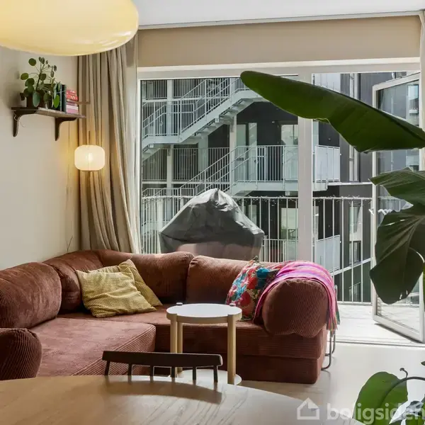 Brun hjørnesofa står i en stue ved siden af et rundt sofabord med puder og tæpper. Store grønne planter ved vinduet afslører en altan med en grill udenfor; bygning strækker sig i baggrunden.