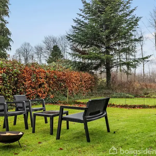 Tre sorte stole og et bord står på en grøn græsplæne. I baggrunden er der høje træer og en hæk med brune blade. Atmosfæren er fredelig og naturlig.