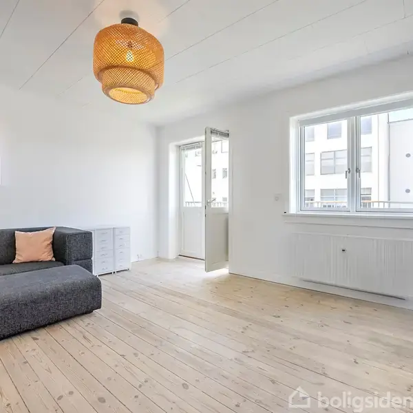 En grå hjørnesofa med en orange pude står i et lyst rum med trægulv. En flettet loftslampe hænger over sofaen, og en dør fører til en balkon.