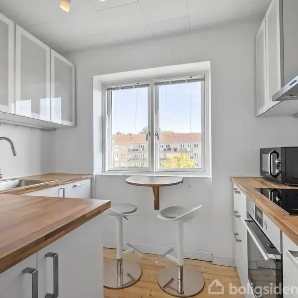 Et lyst køkken med træbordplader og hvide skabe. Et rundt bord med to barstole er placeret ved vinduet, der viser udsigt til naboens bygninger. En sort mikroovn står på bordet.