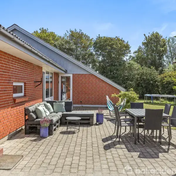 Rød murstenshus med udendørs terrasse. Møbleret med sofa og spisebord med stole. I baggrunden er der en have med græsplæne og træer under en klar blå himmel.