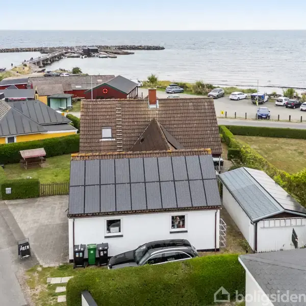 Et hus med solpaneler på taget står tæt ved vandet. Omgivet af andre huse, en båd i en indkørsel og parkerede biler langs vejen ved kysten.