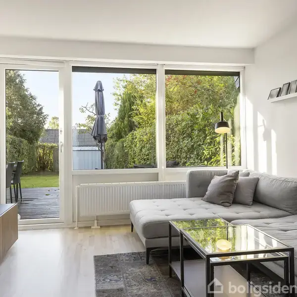 En stor, grå hjørnesofa står i en lys stue med store vinduer, der viser en terrasse og have. Et tv er placeret til venstre. Der er hylder og dekorationer på væggen.