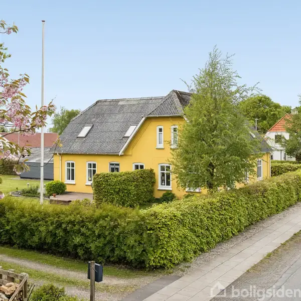 Gul murstenshus med rødt tag af betonfliser omgivet af hække. Det står i en velholdt have ved en stille, grøn villavej med fortov. Blomstrende træer ses i forgrunden.