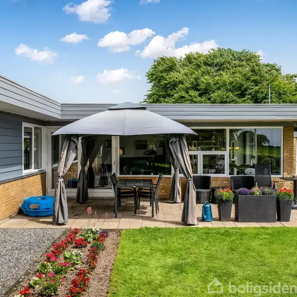 En terrasse med et rundt havepavillon, borde og stole under, omkranset af en moderne bolig med store vinduer og en velplejet have med blomsterbede og grøn græsplæne i solen.