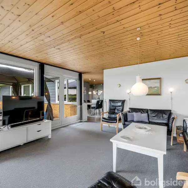 Stue med sofagruppe, fjernsyn på hvidt møbel. To lænestole omkring et sofabord. Store vinduer og dør til terrasse. Træloft og tæppebelagt gulv i moderne boligindretning.