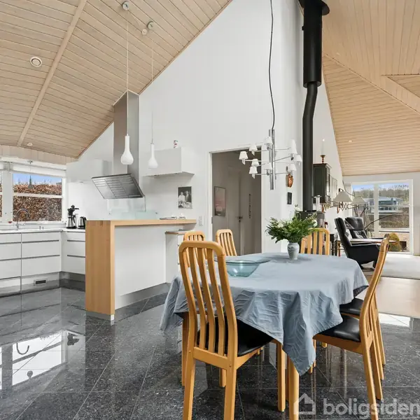 Et moderne køkken-alrum med hvidt interiør og træloft. Et spisebord med blå dug og træstole står i centrum. I baggrunden ses en stue med store vinduer og en brændeovn.