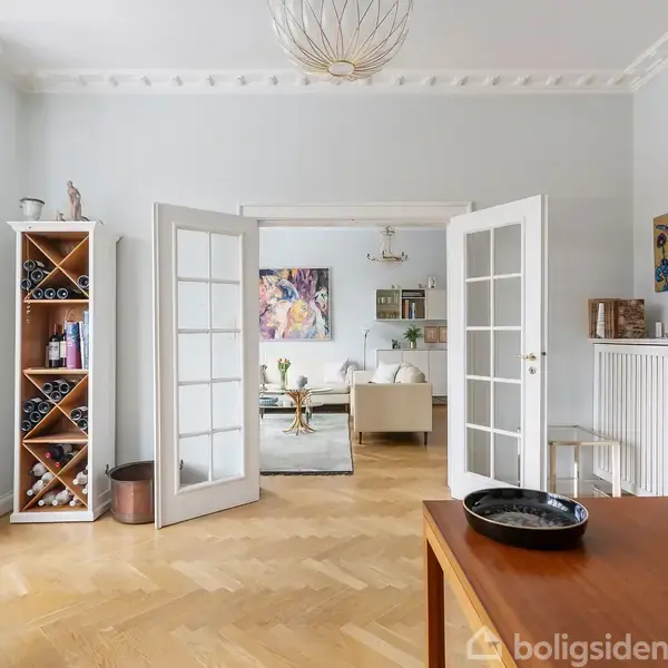 Et lyst rum med parketgulv har franske døre åbne til en stue. En vinreol, kunstværker på væggene, og et træbord med dekoration er på plads.