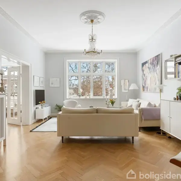 Stue med lys sofa, står stille, i et rum med hvide vægge og trægulv. Store vinduer giver udsigt. Bogreol og kunst på væggene bidrager til en hyggelig atmosfære.