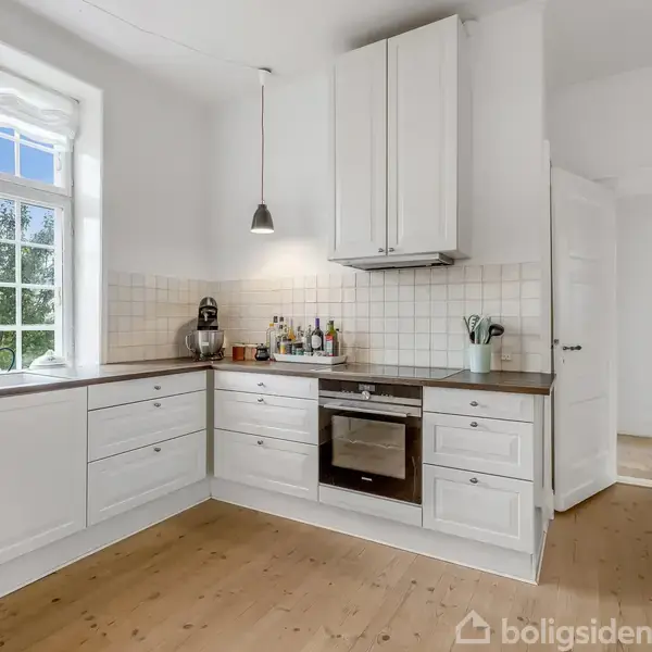 Et lyst køkken med hvide skabe og træbordplade. En ovn er indbygget under kogepladen. Rummet har store vinduer, hængelamper og flisevæg bag ved komfuret.