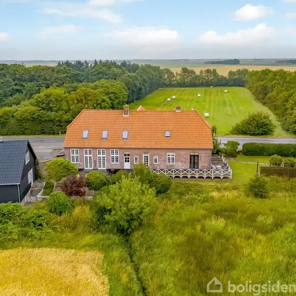 Et stort hus med rødt tag ligger ved en vej, omgivet af grønne marker og skov. I baggrunden ses et stort, åbent landskab.