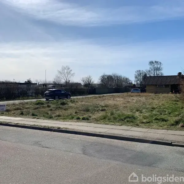 Grunden indeholder et "Til salg" skilt og bil holder i baggrunden. Den er omgivet af huse og træer på en rolig vej under en blå himmel.