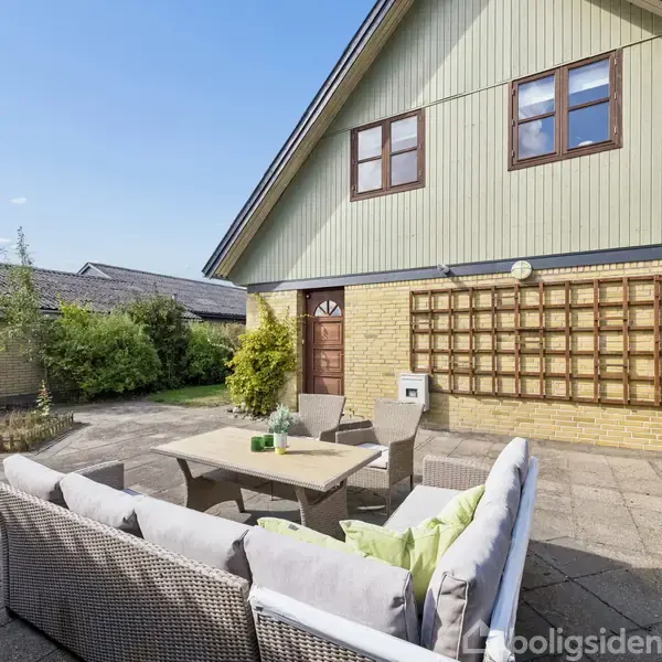Rattan sofa med hynder står på en flisebelagt terrasse foran et gulstenshus med grønt træværk. Et lille bord med glas står ved siden af, omgivet af buske.