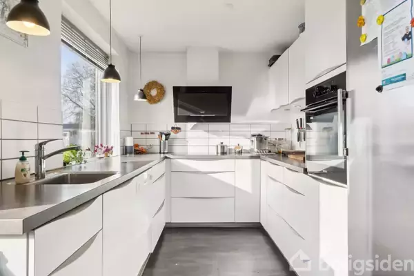 Et moderne køkken med hvide skabe og stålbordplader; indeholder en ovn, kogeplade og vask. I lyse omgivelser med hængelamper og udsigt til natur gennem et stort vindue.