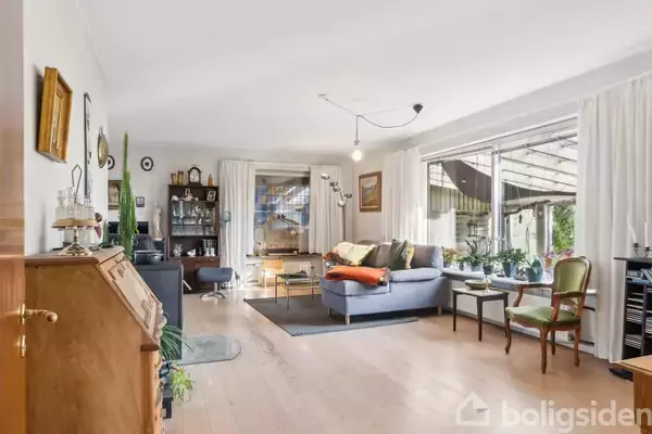 En stue indrettet med sofa, puder og lænestole. Store vinduer giver naturligt lys, mens gardiner hænger ved siden af. Flere møbler og dekorationer er til stede i en hyggelig atmosfære.