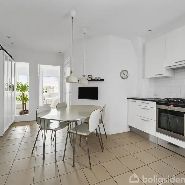 Køkken med et rundt bord og fire stole, omgivet af hvide skabe og en stålkomfur. En plante står ved glasdøre, og et ur hænger på væggen med fliser.