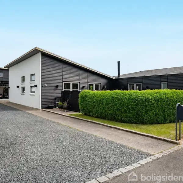 Et moderne hus med sort og hvid facade står i en velplejet have med grusindkørsel. En grøn hæk omkranser ejendommen. Blå himmel ses i baggrunden.