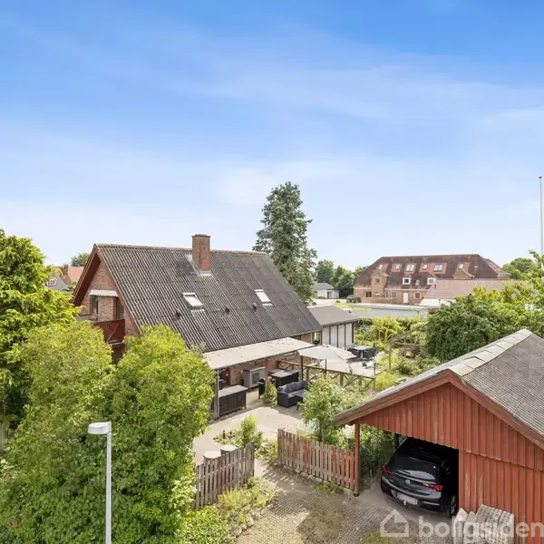 Et rødt hus med sort tag ligger omgivet af grønne træer og en carport til højre. Huset er i en rolig, beboelig område med flere bygninger i baggrund.
