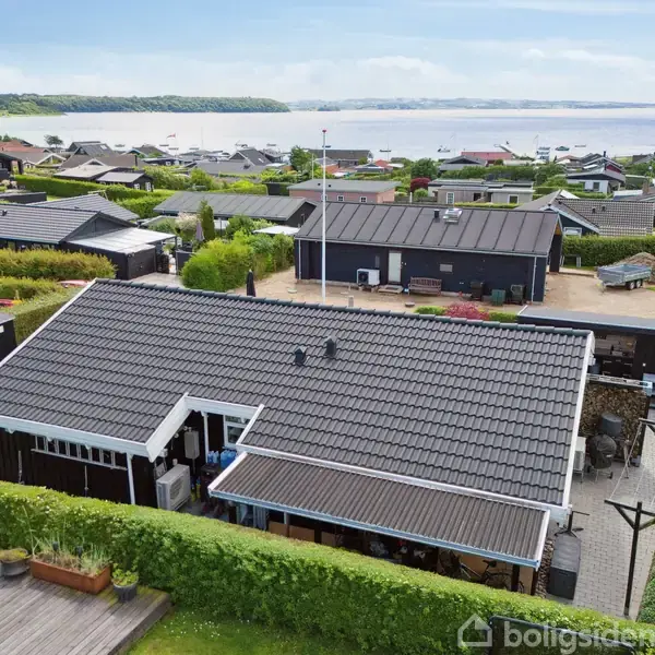 Et sort hus med gråt tag, omgivet af hække og træterrasse, ligger i et beboelsesområde nær en kystlinje med udsigt til vandet og små både i det fjerne.