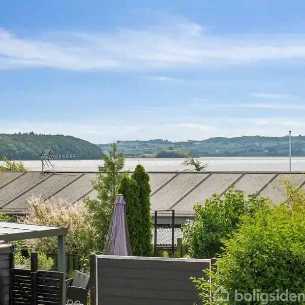 Renoverede tage ses forgrunden med en landovergang i baggrunden. Havudsigt fylder horisonten, omgivet af frodige buske og træer under en klar blå himmel.